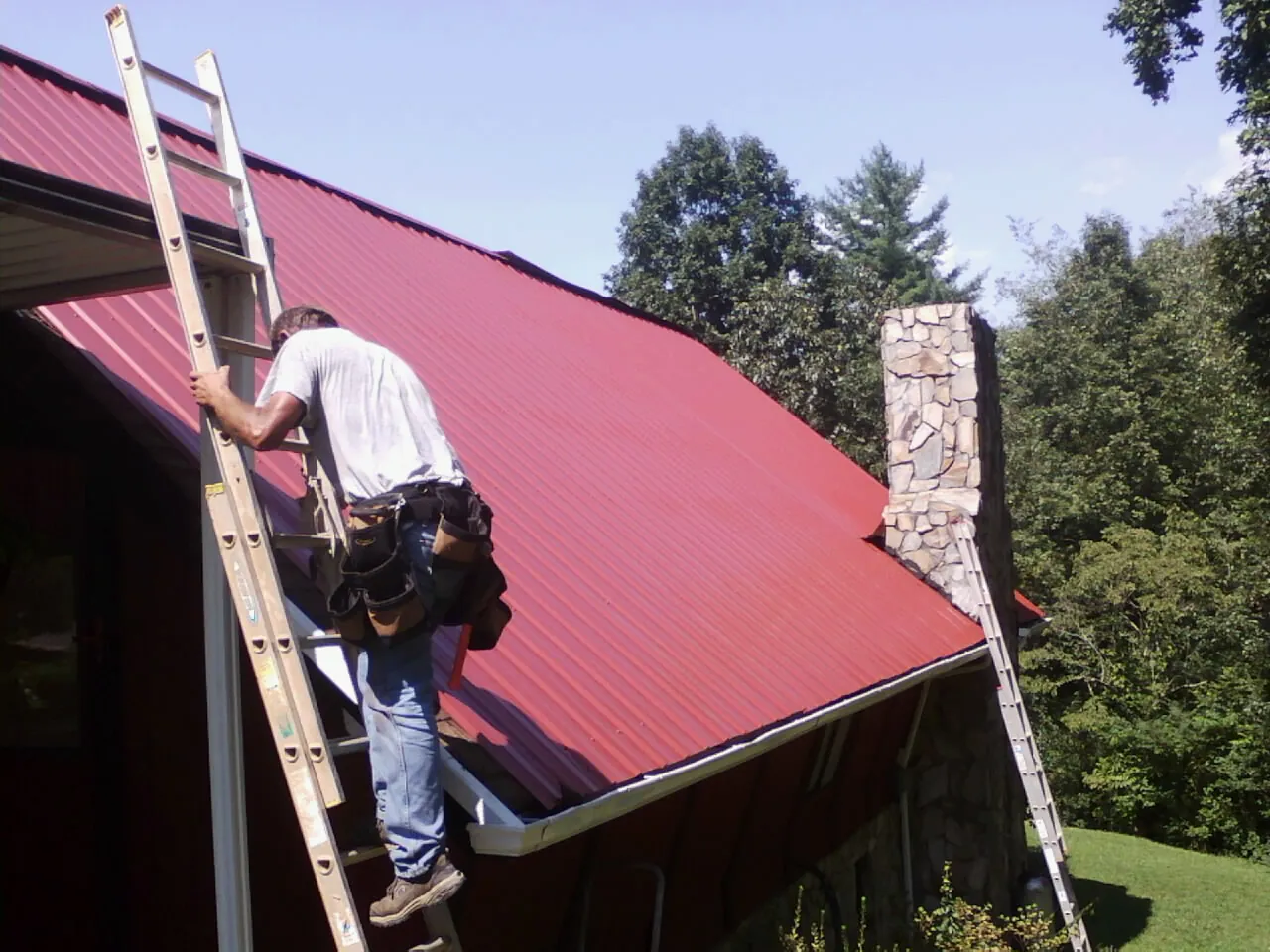 Roofer on a ladder beside a red metal roof and stone chimney — Soffit & Fascia Repair by Roofing Brothers in Coventry